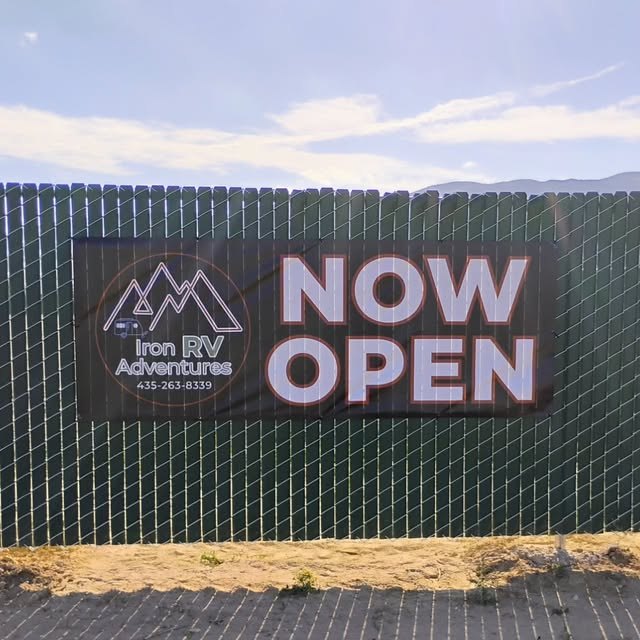 The Iron RV Adventures “Now Open” banner hung on the privacy-slatted fence around the storage yard, mountains behind.
